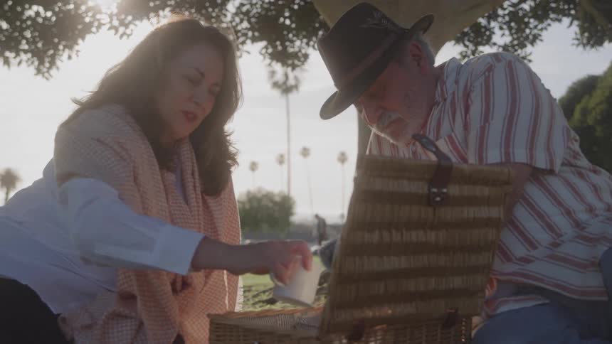 An elderly couple grabbing mugs from a wooden basket while sitting on a park on a sunny day