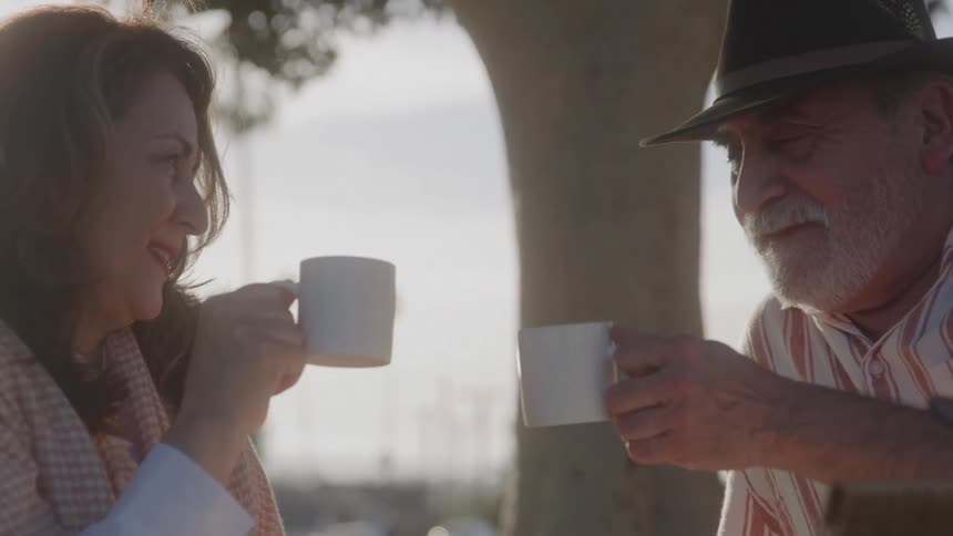 Two people sit facing each other with mugs in their hands the people move the mugs toward each other and clink them together