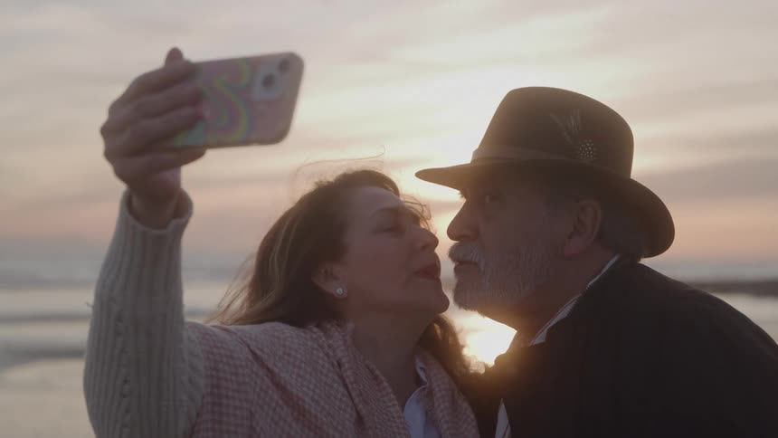 A man and a woman take a selfie with a phone while kissing in front of a sunset over the ocean