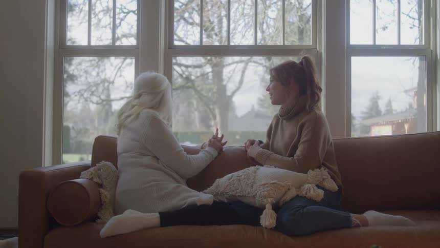Two women are having a conversation on a couch next to a large window