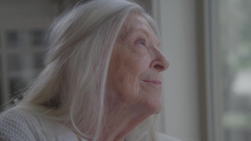 Elderly woman with white hair smiles softly while looking out the window