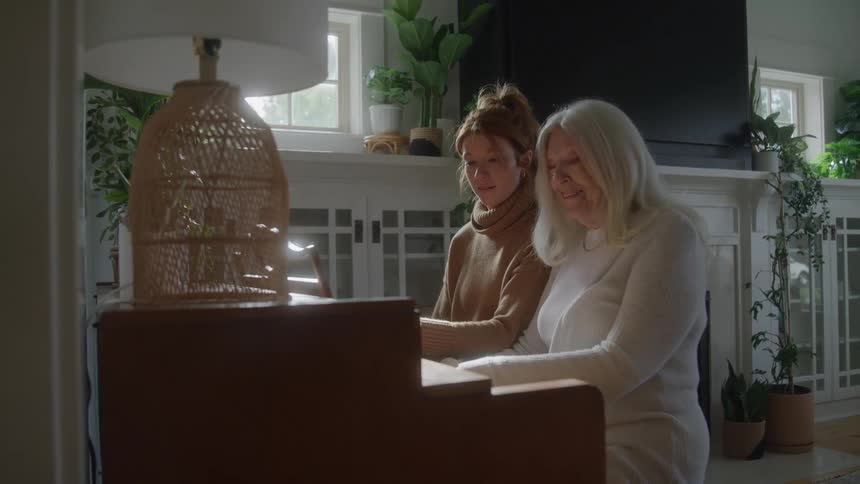 A young woman and an elderly woman are sitting together on a piano bench with the younger woman teaching the older woman to play