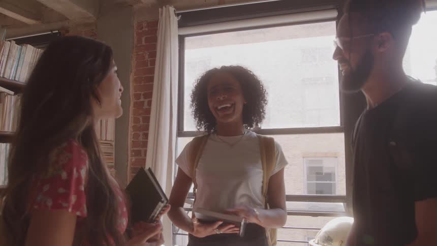 Three people stand in a library setting next to a window and talk