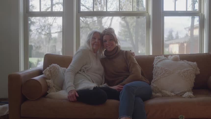Elderly woman and young woman sitting together on a brown couch smiling and hugging in front of a large sunlit window