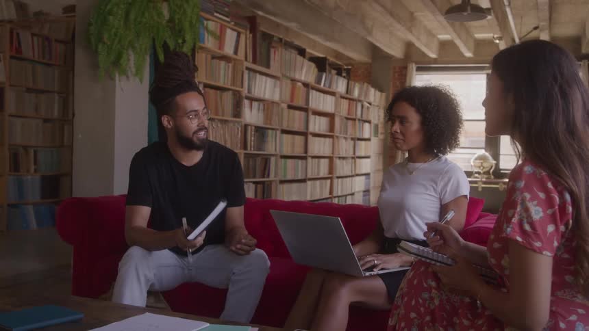 Three casually dressed adults two women and one man are sitting on a red couch with notebooks and a laptop discussing a project in a loft with tall bookshelves lining the wall