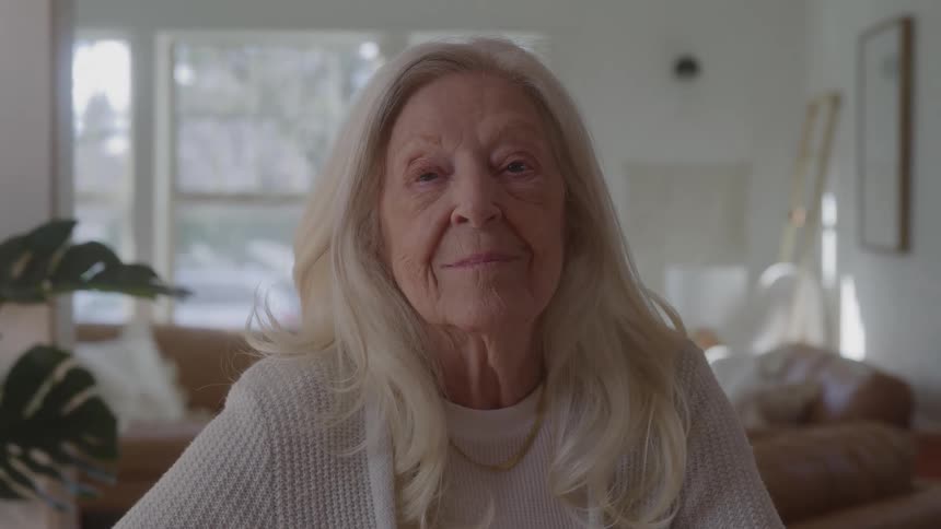 Elderly person with light hair wears white sweater and gold necklace in room lit by large windows with modern and cozy decor