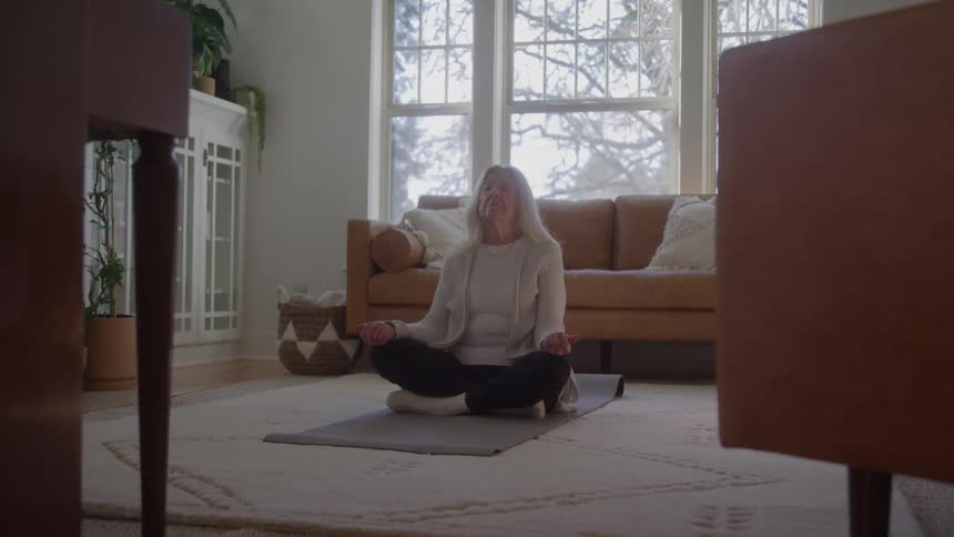 Person meditates seated crosslegged on mat in room lit by natural light with lightcolored furniture and decorative plants