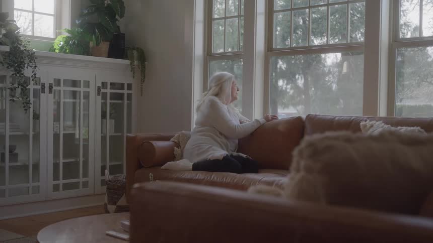 A woman with gray hair sits on a couch in a living room and looks out a window