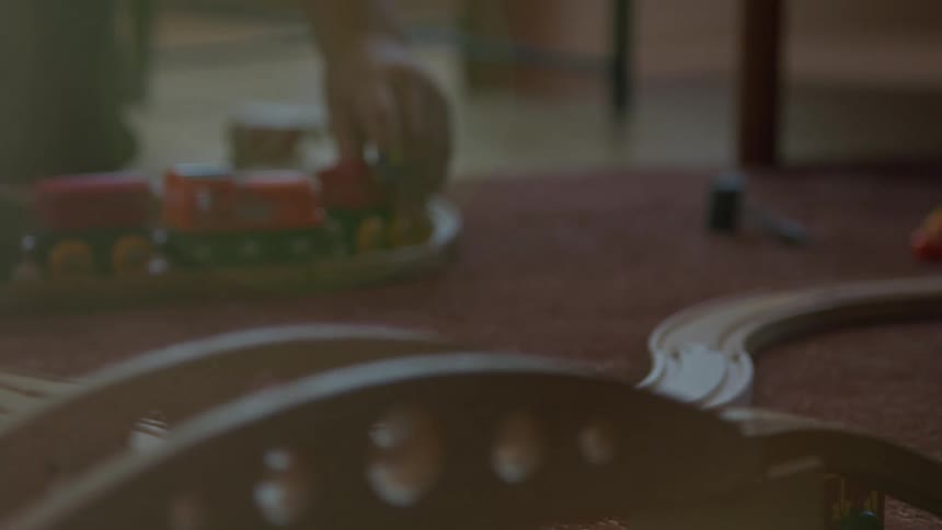A boys hand pushes a toy train on a wooden track across a floor next to a bridge