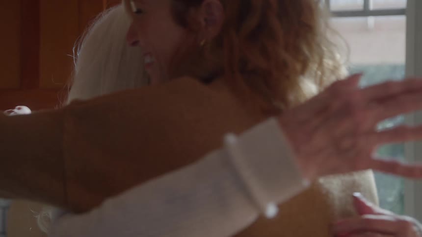 An elderly woman smiling as she hugs a young woman wearing glasses in the kitchen at midday