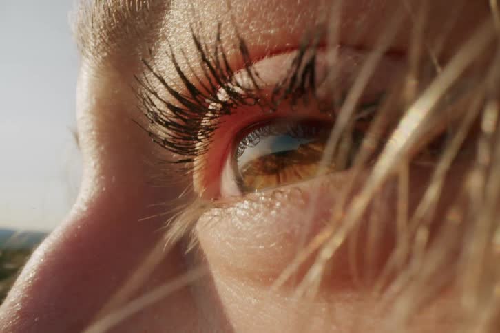 A close view of a womans eye with long dark eyelashes shows a blurred reflection of a bright sunlit landscape with a blue sky