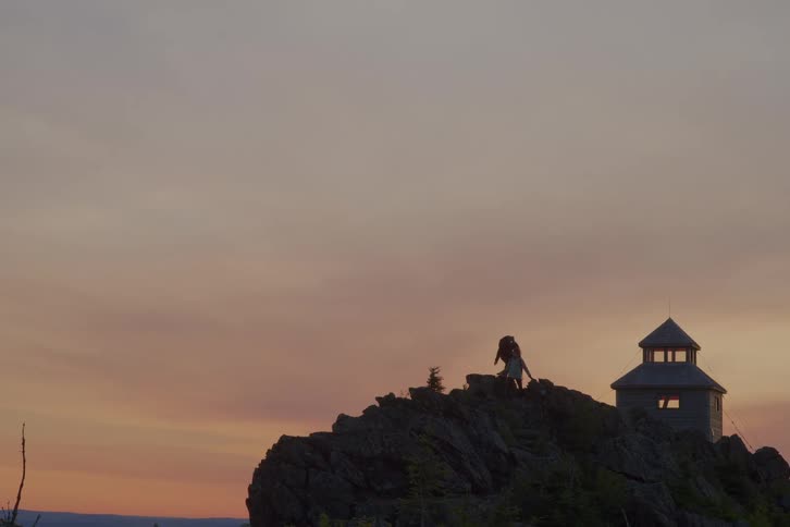 Three people hiking on the peak of a mountain and a building with a tower is on the background and theres a cloudy sky on the sunset