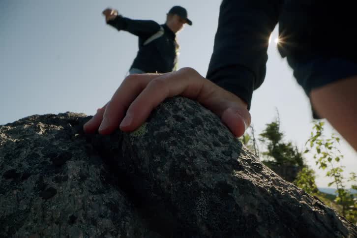 Two people in shorts and dark jackets are climbing over a large rough rock formation with the bright sun flaring in the background