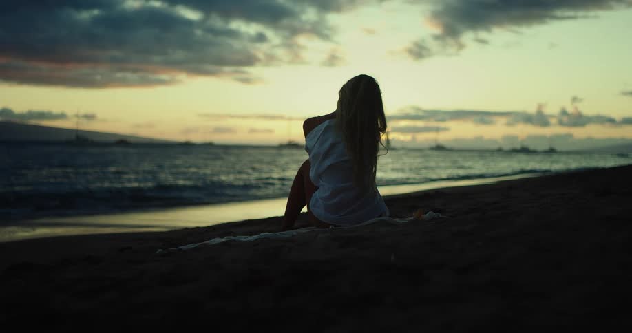 A woman with long blonde hair sits on the beach looking out at the ocean as the sun sets the water gently laps at the shore as the sky glows in a mix of orange and red