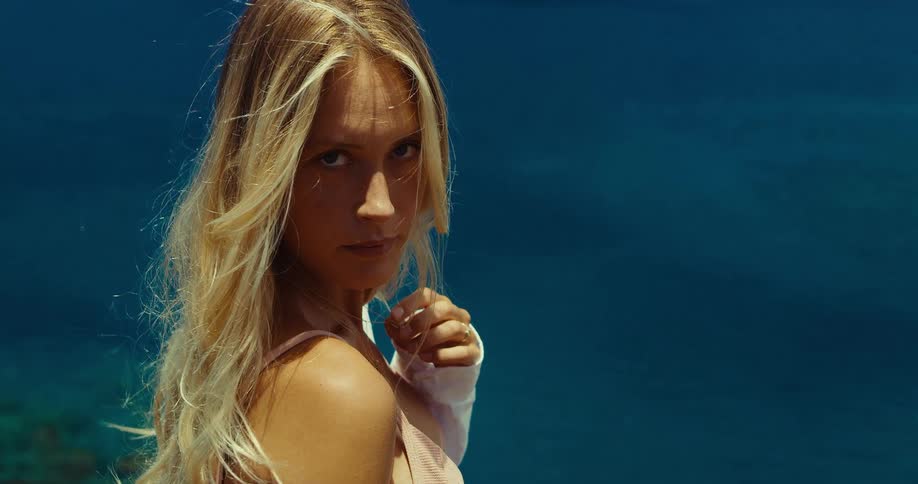 A young woman with long blonde hair looks directly at the camera with a serious expression shes wearing a lightcolored top and a deep blue background is visible behind her