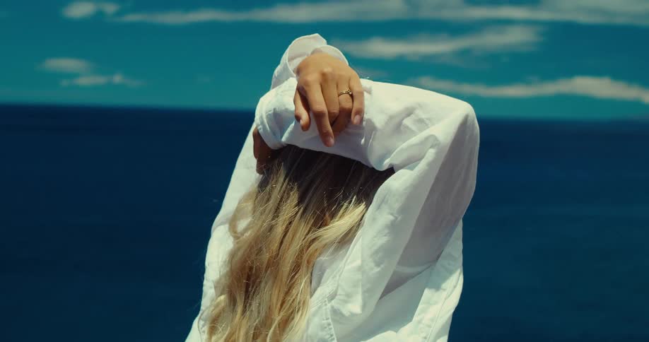 Blonde woman wearing a white shirt stands on the coast looking at the sea with her arms resting on her head