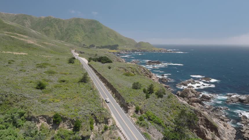 Vehicles drive along a highway on a coast with a steep drop on a sunny summer day