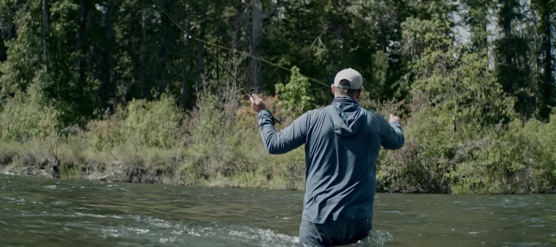 A man in a hat and a longsleeved shirt is standing in the middle of a river fly fishing he is surrounded by trees on the riverbanks