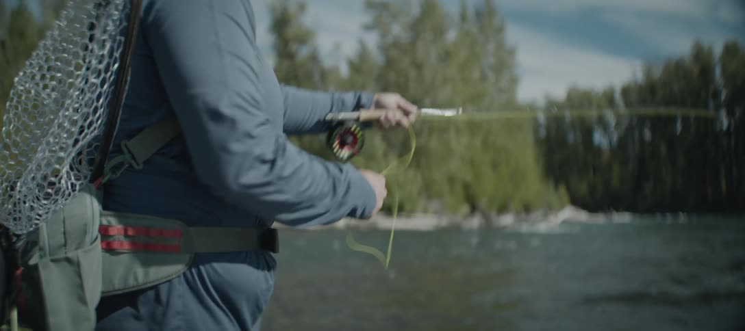 A man stands and holds a fishing rod the mans hands move as they cast a line into the water