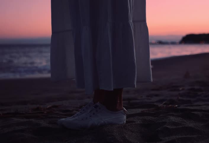 A woman standing on a beach in the sunset wearing sneakers and a white dress