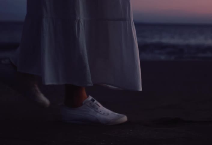 A woman in a white dress and white sneakers walks along a dark sandy beach as gentle waves roll in at dusk