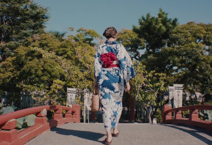 A woman in a vibrant blue and white floral kimono with a red obi walks away from the camera across a traditional red bridge
