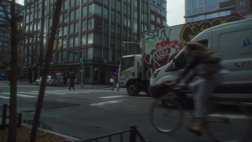 A bicyclist rides quickly across a busy city street intersection in front of a white box truck covered in red graffiti and a yellow school bus surrounded by tall buildings on a cloudy day