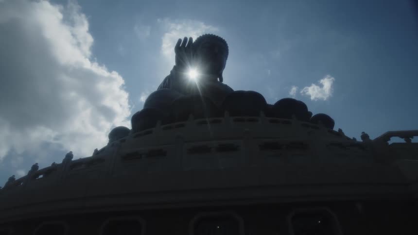 Sunlight shines between the hand and the neck of a large statue of buddha sitting on a building on a cloudy day