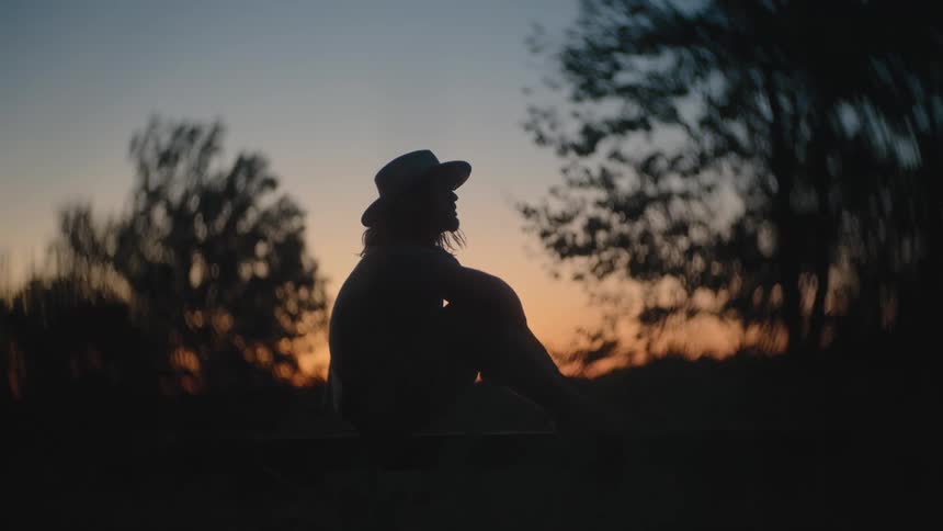 Person wearing widebrimmed hat sits outdoors at sunset with trees in the background