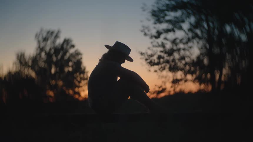 A person sits alone outdoors silhouetted against a setting sun with trees in the background