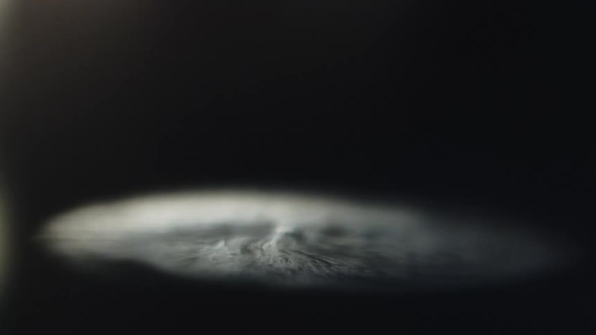 A single spotlight illuminates the surface of a dark liquid as a puff of white smoke or dust appears and then slowly dissipates