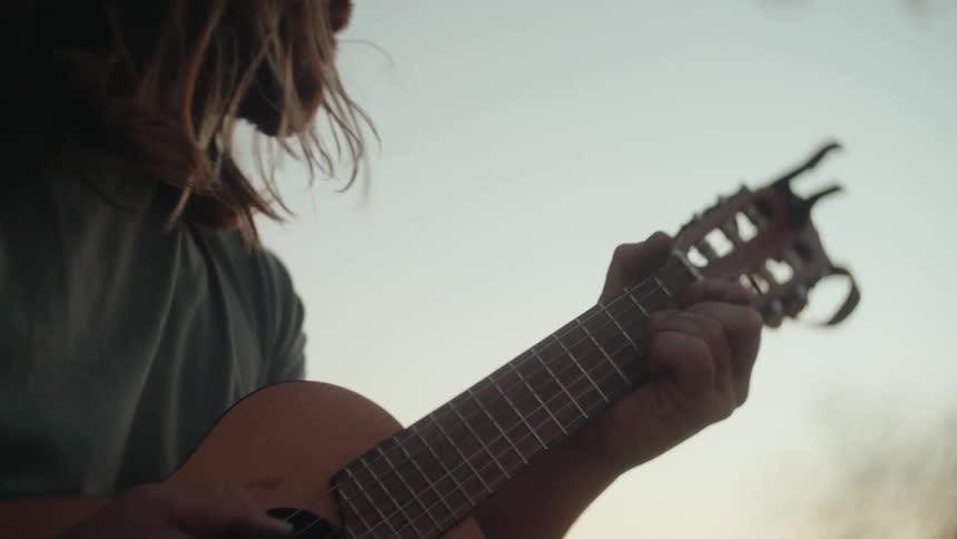 A young man plays a guitar in an outdoor setting the mans hands move along the guitars neck and strings