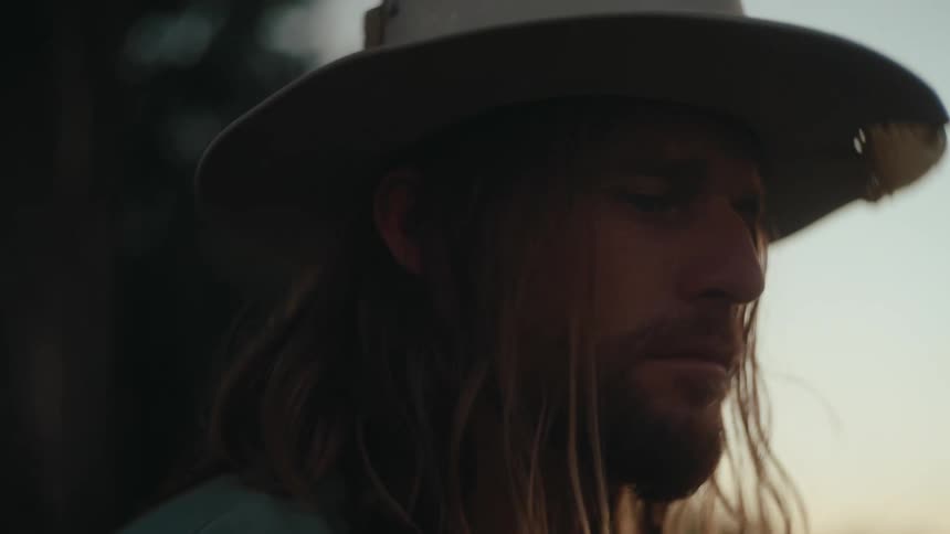 A man wearing a hat with long hair and beard nodding his head as he plays guitar in a field at dusk