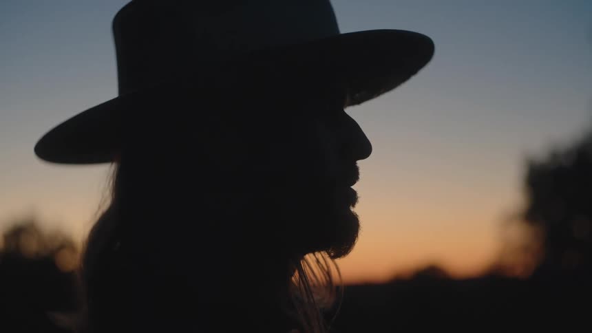 A man wearing a hat stands silhouetted at sunset