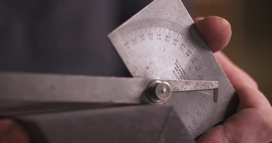 Mechanical protractor with 0 to 180 degree scale is held by hand in front of blurred metallic surface