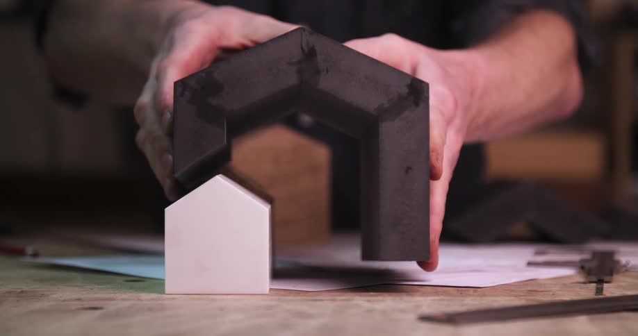 Hands position a dark structure over a small white house model