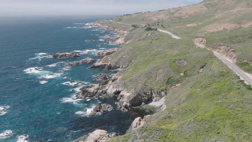 The ocean waves crash onto a rocky shoreline at the base of large grassy hills