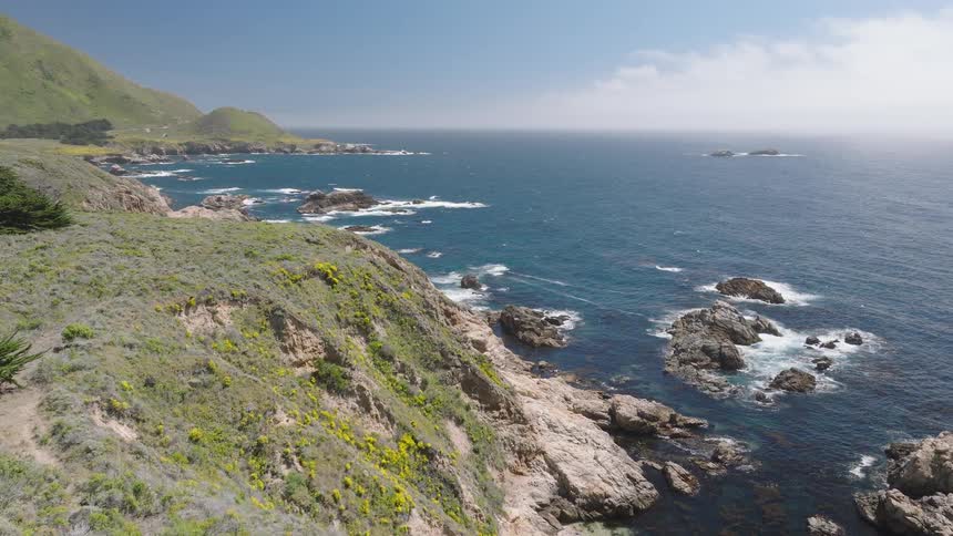 An aerial shot captures a scenic view of a vast blue ocean with waves crashing against a rocky coastline the cliffs are covered in green and yellow vegetation