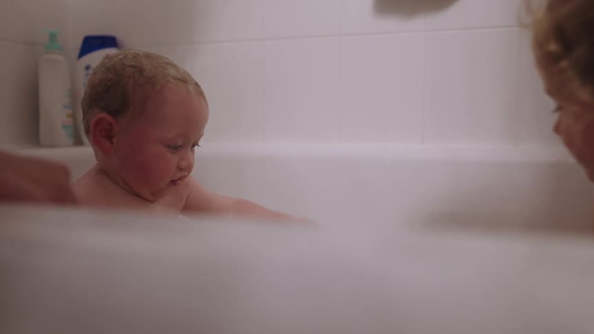 A mother bathing a female toddler and an infant in a bathtub at midday with the baby joyfully splashing and smiling during bath time