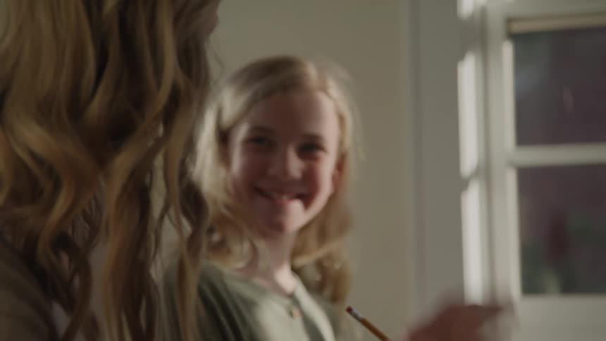 A smiling girl wearing a green cardigan finishes writing with a pencil then high fives an older woman sitting beside her who is assisting with homework