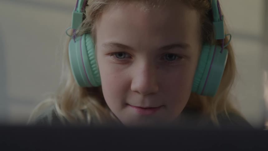 A close view shows a young woman with blonde hair wearing bright turquoise headphones smiling slightly while looking intently at a laptop screen
