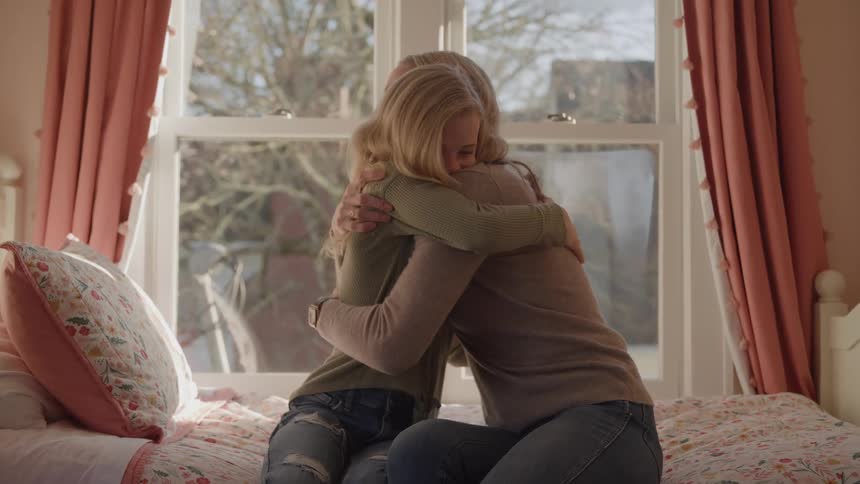 Two woman sit on a bed and embrace in a hug