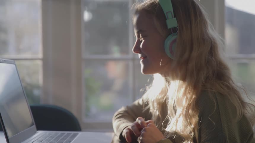 A young woman wearing bright turquoise headphones smiles and waves at the screen of a laptop while sitting near a sunlit window