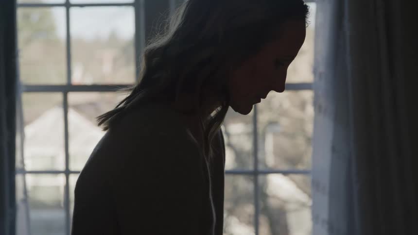 A profile silhouette of a woman with long hair is seen against a bright window frame looking downward with a thoughtful or sad expression