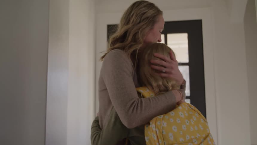 A smiling woman wearing a brown sweater and jeans helps a young woman put on a yellow patterned backpack gives her a hug and watches her walk out the front door