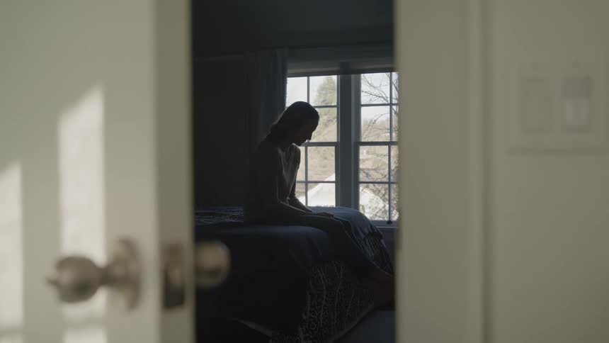 A young woman sits on a bed and looks down in a bedroom setting