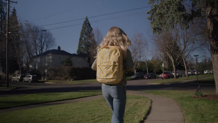 A girl with long blonde hair wearing a bright yellow patterned backpack and blue jeans walks away on a winding sidewalk in a sunny tree lined suburban neighborhood