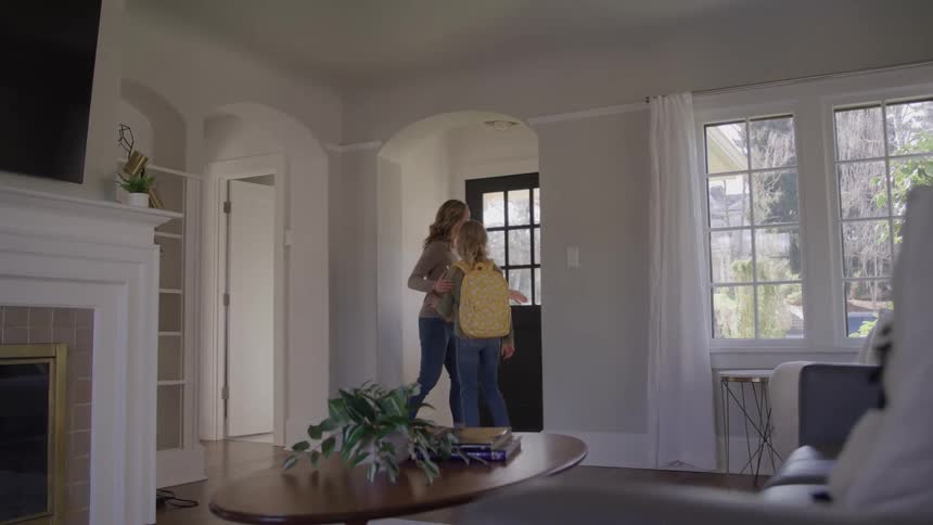 A woman helps a girl adjust a backpack in a living room near a front door the two embrace and the girl then turns and walks out of the house through the open doorway