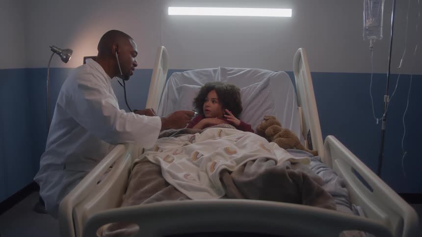 A bald man wearing a white doctors coat uses a stethoscope to examine a young girl lying in a hospital bed with a teddy bear and an iv drip visible nearby