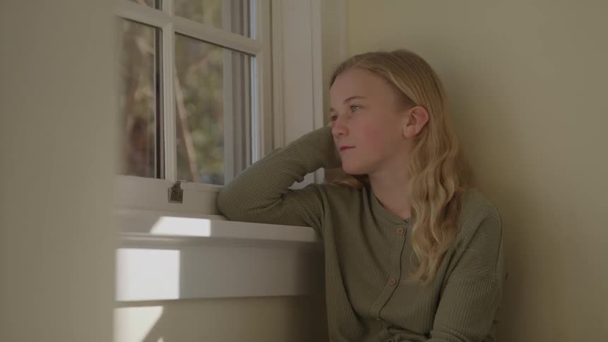 Blondehaired teenager looks out the window with head resting on hand in bright indoor setting